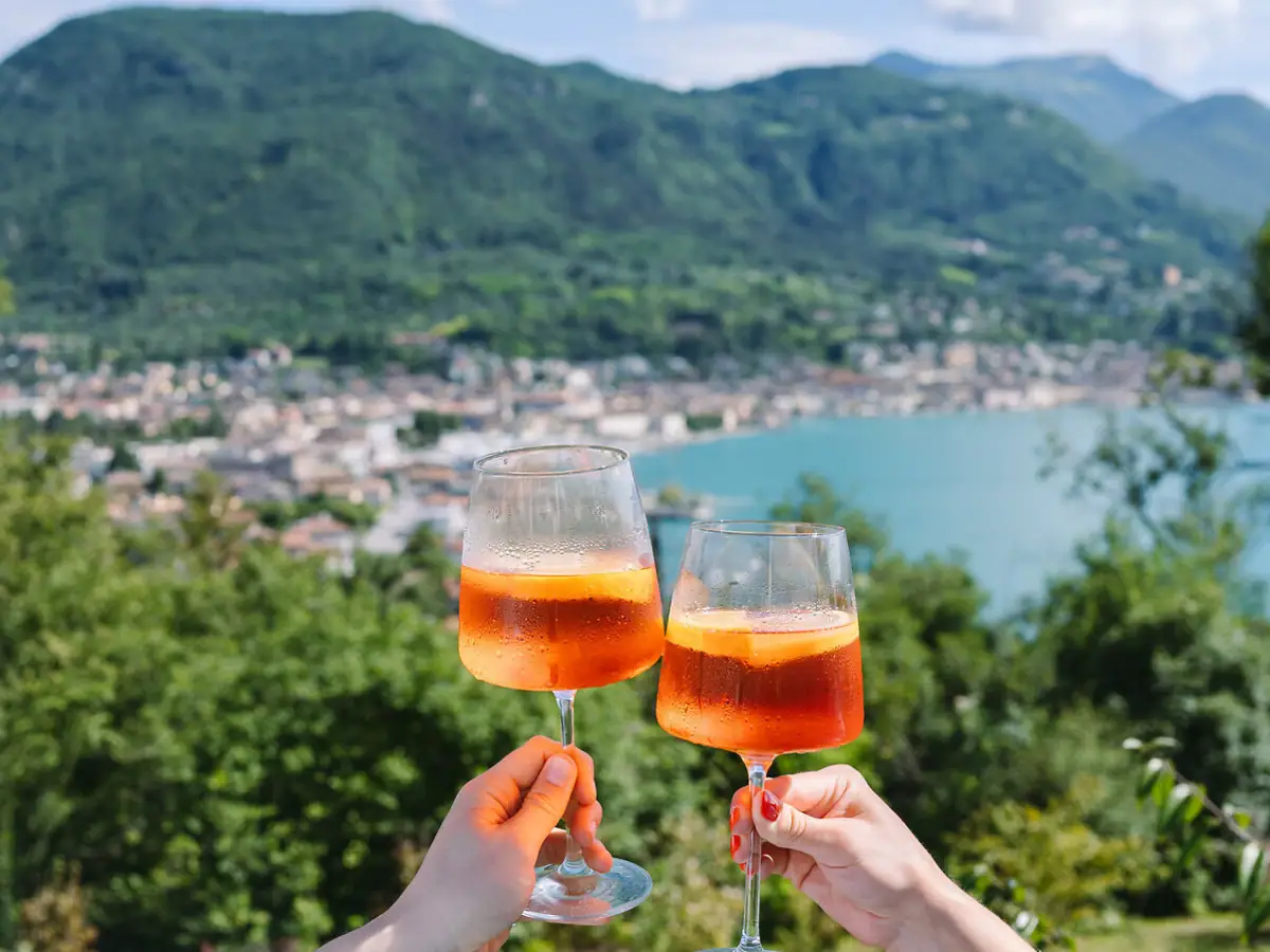 Zwei Gläser Aperol Spritz werden über dem Gardasee angestoßen, im Hintergrund das Panorama der italienischen Berglandschaft.