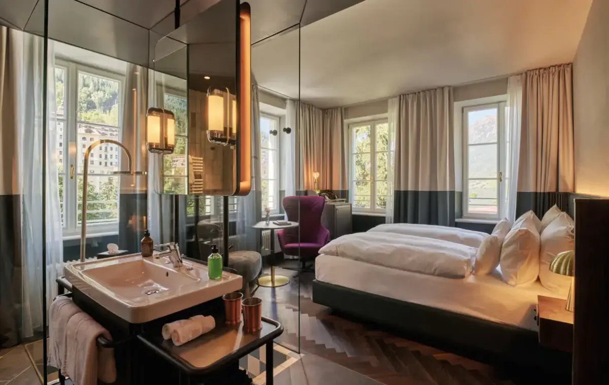 Blick in ein Hotelzimmer mit verglastem Badbereich, Doppelbett und Sitzgelegenheit am Fenster.