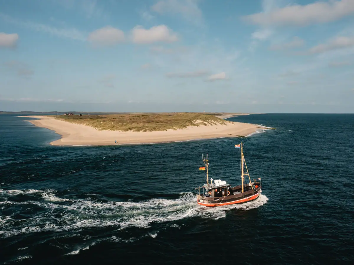 Luftaufnahme eines traditionellen Fischerboots auf offener See nahe einer malerischen Sandbank unter einem klaren blauen Himmel. Die Szene strahlt Ruhe und Abgeschiedenheit aus, während das Boot sachte Wellen auf dem Wasser hinterlässt und sich seinen Weg durch das Meer bahnt. Im Hintergrund ist die weite, unberührte Landschaft zu erkennen, die die Naturschönheit der Küstenregion hervorhebt.