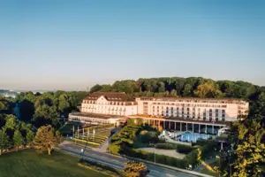 Großes weißes Gebäude mit Pool im Vordergrund und Bäumen im Hintergrund.