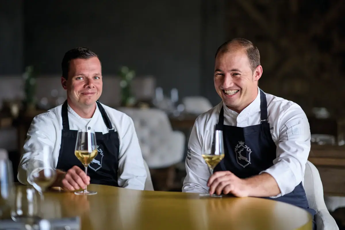 Hans und Sascha am Messingtisch im Ifen Hotel Zwei Männer in Schürzen sitzen an einem Tisch mit Weingläsern.
