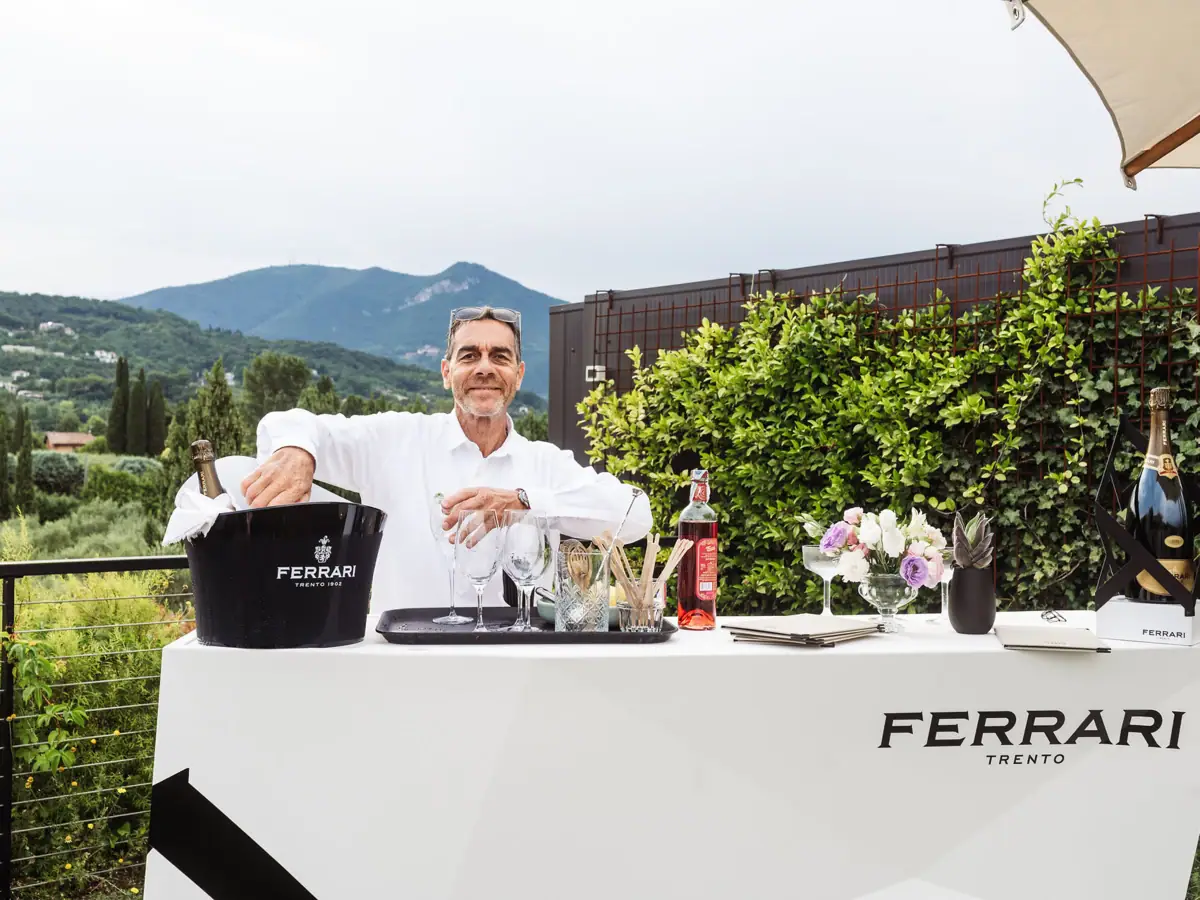 Barkeeper bereitet an einem Ferrari-Trento-Tisch Cocktails zu, umgeben von mediterraner Natur und Bergblick.