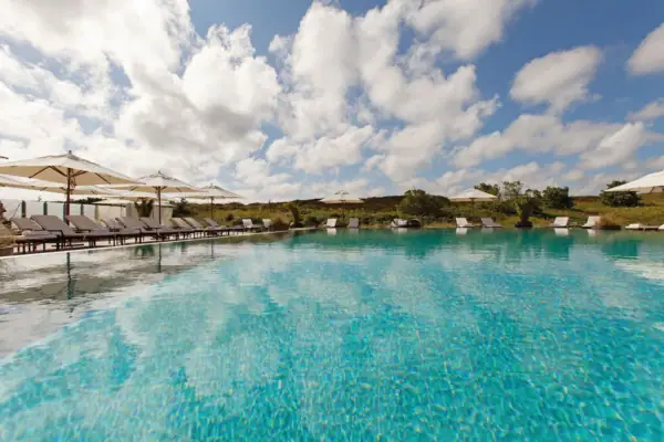 Ein Pool mit Liegestühlen und Sonnenschirmen und im Hintergrund sind Dünen zu sehen.