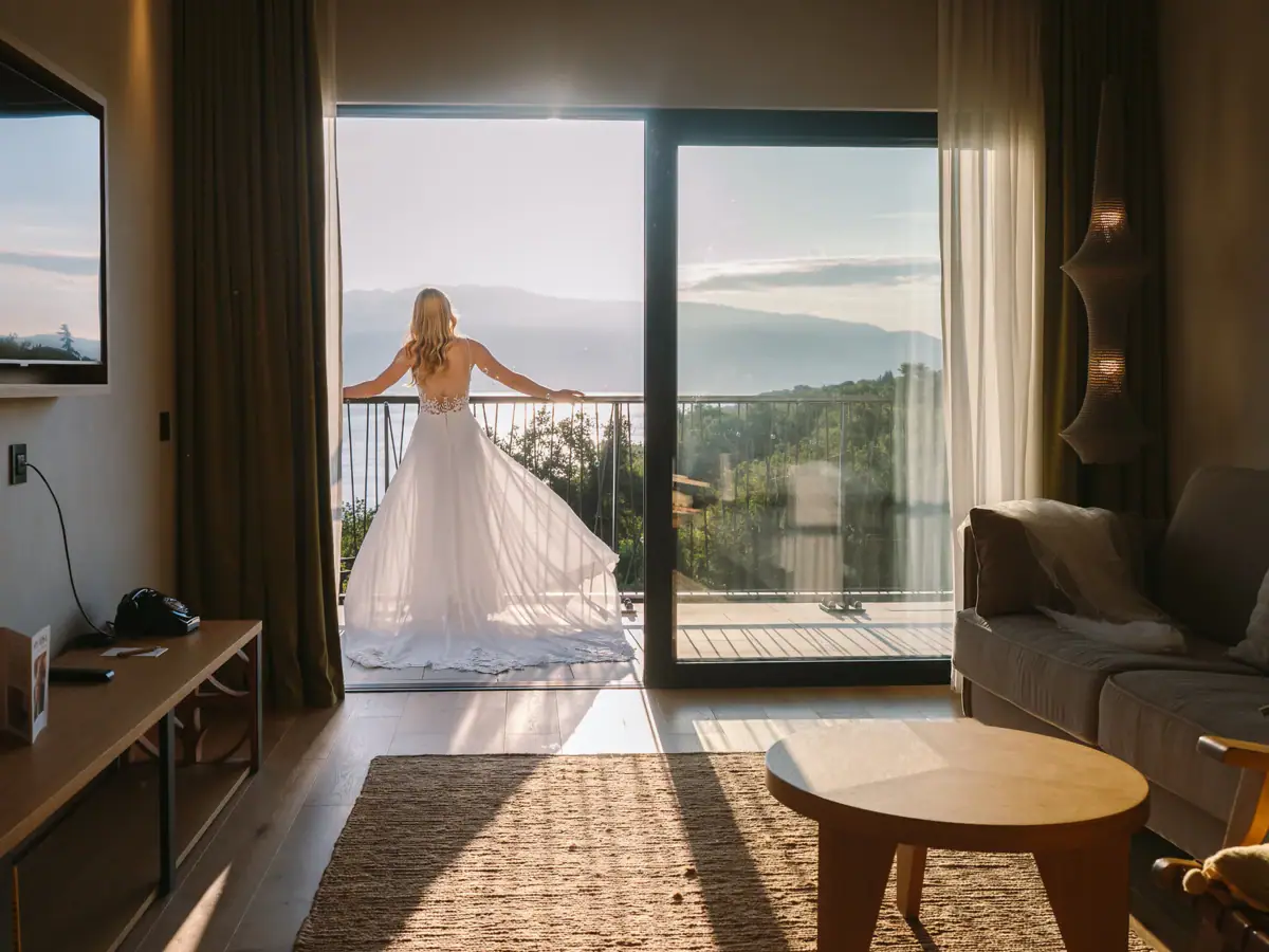 Braut öffnet die Balkontüren ihrer Suite und blickt im Hochzeitskleid auf den Gardasee.