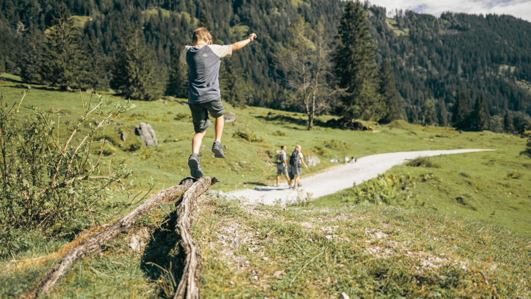Ein Kind in Wanderkleidung springt über eine Wurzel in den Bergen. Im Hintergrund sieht man die Familie auf einem Wanderweg.
