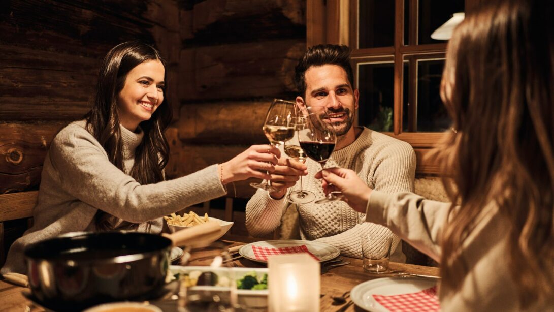 Zwei Frauen und ein Mann stoßen mit Wein zum Essen an. Auf dem Tisch stehen Teller, eine Kerze und ein Topf.
