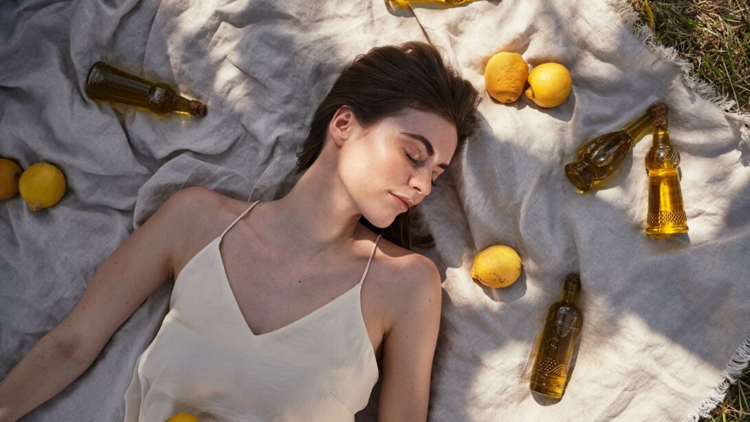 Eine Frau liegt auf eine Decke mit Zitronen und Olivenölflaschen. Licht scheint auf ihr Gesicht.