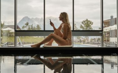 Frau im beigen Bikini sitzt mit Handy am Pool auf einem Handtuch vor einer großen Fensterfront