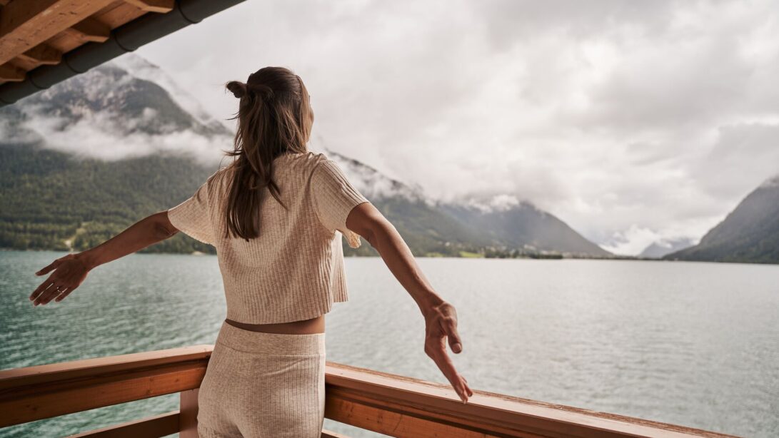 Eine Frau steht auf dem Balkon und schaut mit zur Seite geöffneten Armen auf den Achensee