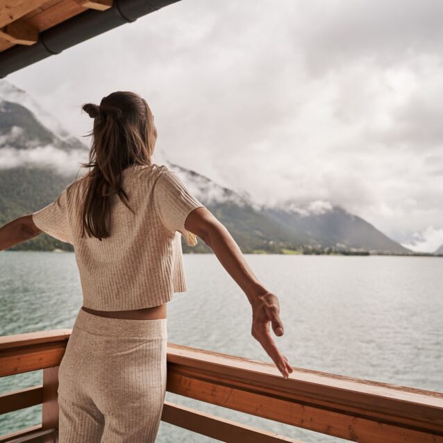 Eine Frau steht auf dem Balkon und schaut mit zur Seite geöffneten Armen auf den Achensee