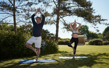 Eine Frau und ein Mann stehen in der Yogaposition "Baum" auf einer Yogamatte in einem grünen Garten auf einer Rasenfläche. Der Schatten eines Baumes schützt Sie vor der Sonne.