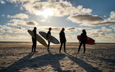 Vier junge Menschen im Neoprenanzug stehen an einem Strand. Drei von ihnen haben Surfbretter in der Hand. Die Sonne wirft ihren Schatten in den Sand.