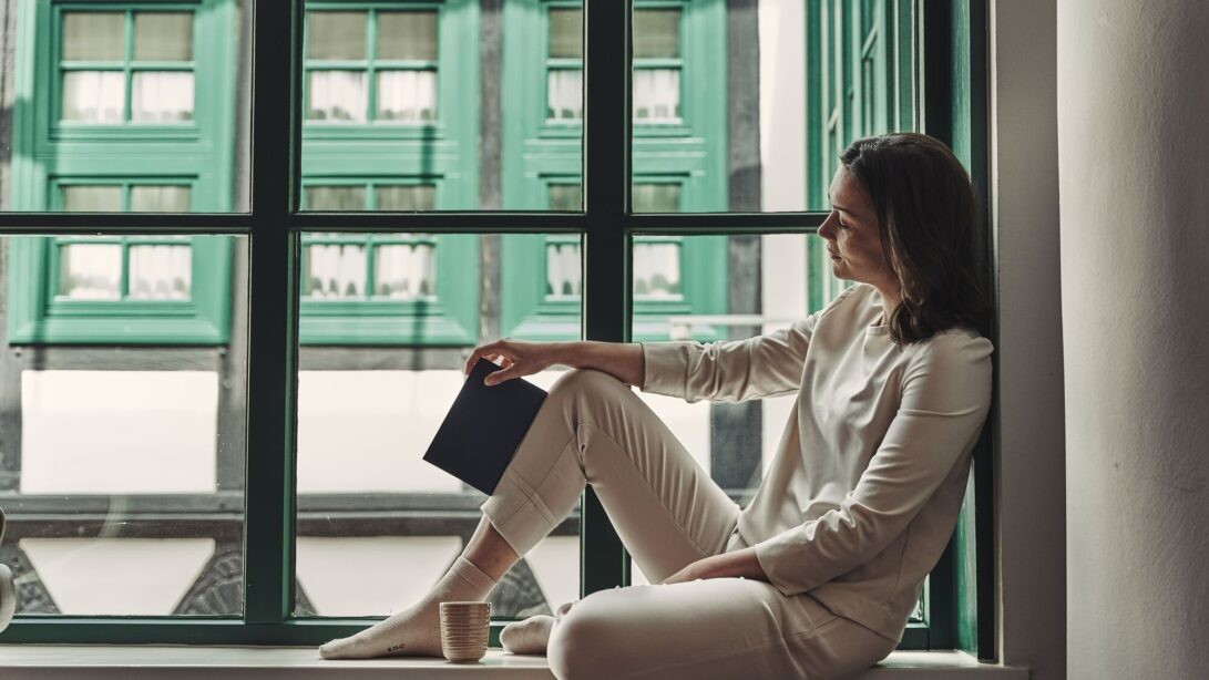 Eine Frau sitzt in Loungewear auf der Fensterbank. Sie hält ein geschlossenes Buch in ihrer rechten Hand und eine Tasse steht vor ihr. Im Hintergrund erkennt man eine historisch anmutende Fassade.