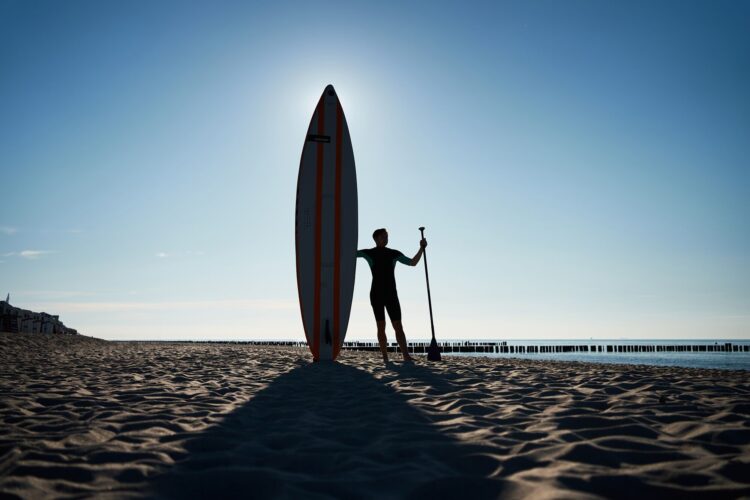 Ein Mann im Neoprenanzug mit SUP und Paddel in der Hand steht an einem Strand und wird von hinten von der Sonne angeschienen. 