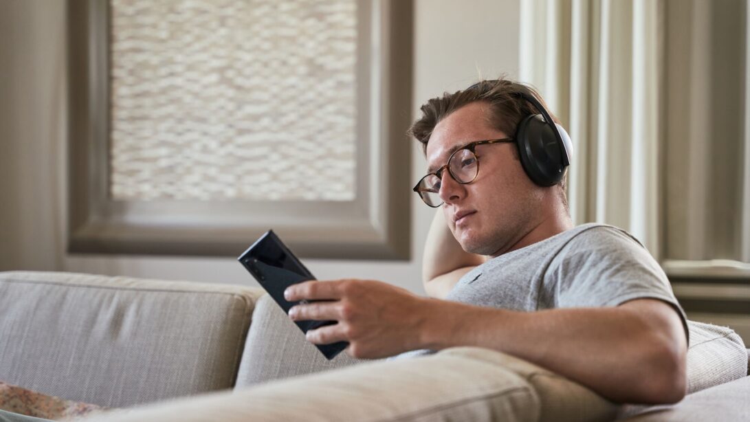 Mann hört Podcast auf Sofa mit Kopfhörern und schaut auf sein Handy