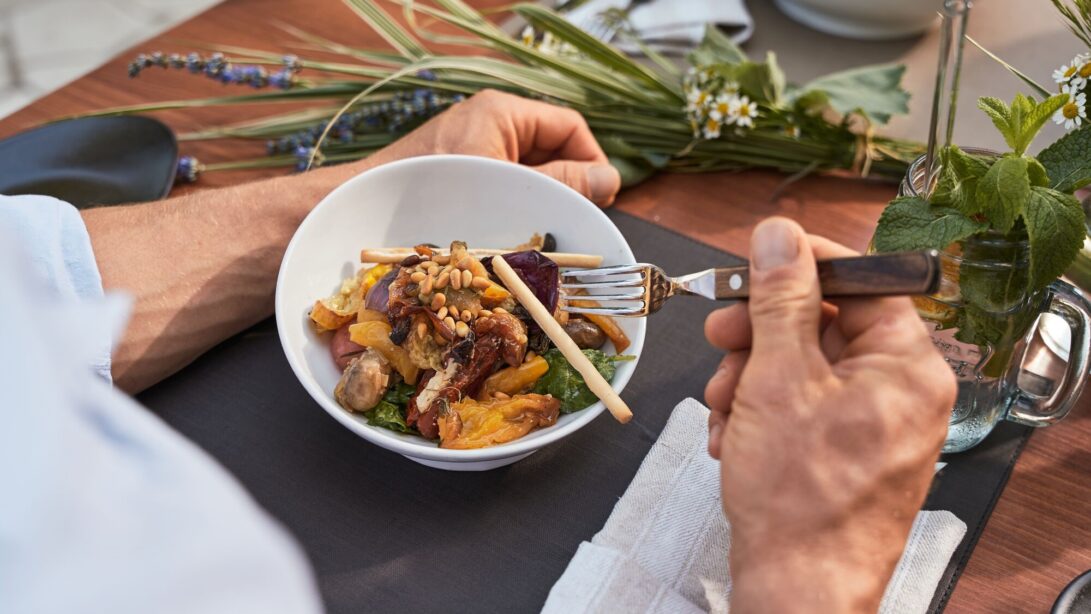 Männerhände halten eine Gabel in Richtung einer Schüssel mit gebratenen Gemüse, daneben steht ein Getränk mit Minze und Zitrone und Wildblumen liegen auf dem Tisch