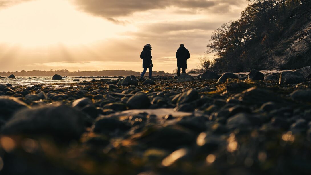 Zwei Personen spazieren bei Dämmerung an einem steinigen Strand an der Ostsee