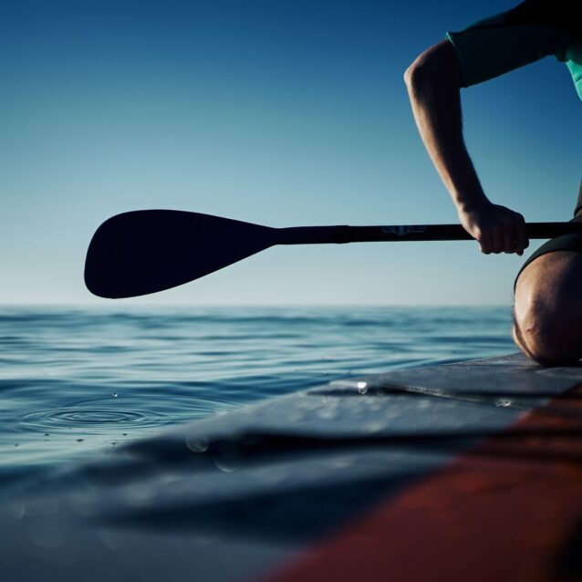 Ein Mann kniet auf einem Stand up Paddleboard im Wasser und hält das Paddel fest. Tropfen fallen vom Paddle ins Wasser.