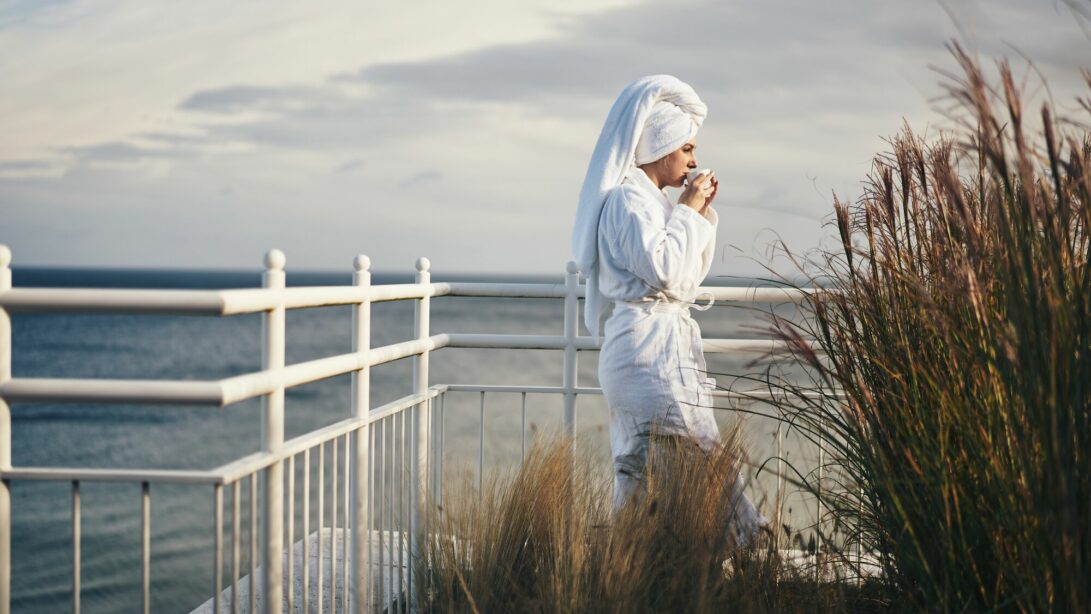 Frau Steht im Bademantel und mit Kopftuch im Haar auf Terrasse. Im Hintergrund das Meer.