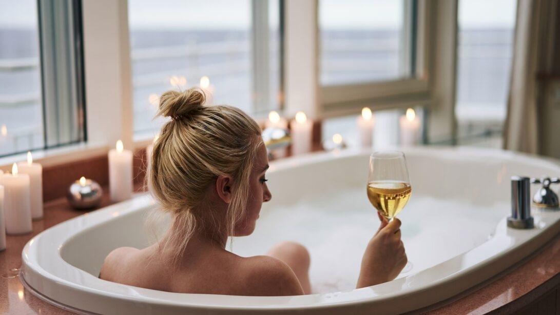 Eine Blonde Frau sitzt mit Weinglas in einer Badewanne welche von Kerzen umringt ist. Im Hintergrund ist eine Fensterfront mit Blick in die Ferne.
