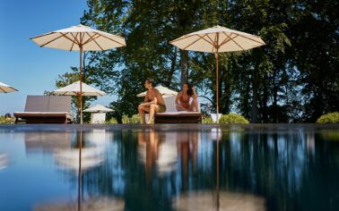 Mann und Frau sitzen in Badebekleidung auf einer Doppelsonnenliege im Schatten eines Sonnenschirms vor einem Pool, welcher das Motiv verschwommen spiegelt