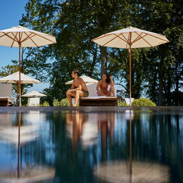 Mann und Frau sitzen in Badebekleidung auf einer Doppelsonnenliege im Schatten eines Sonnenschirms vor einem Pool, welcher das Motiv verschwommen spiegelt
