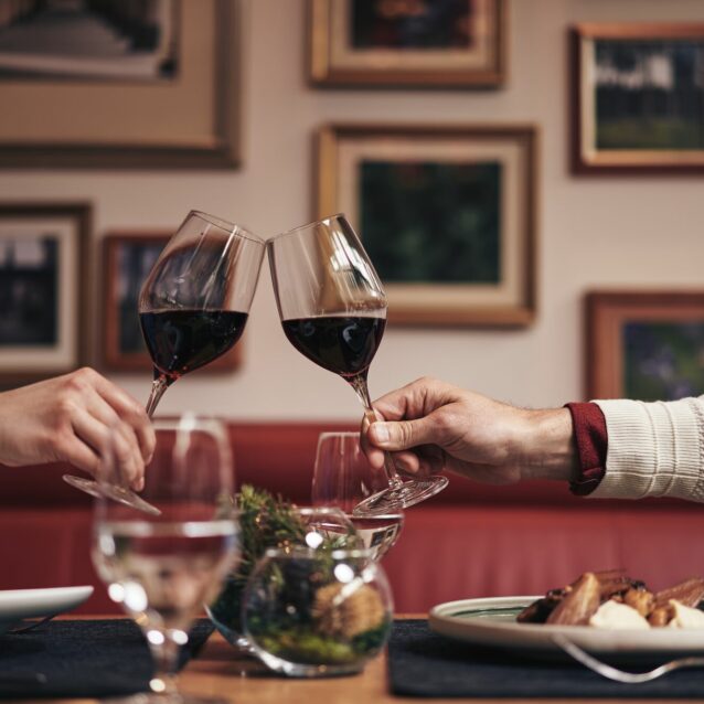 Zwei Weingläser stoßen an, welche von jeweils einer Hand von zwei verschieden Personen über einem Tisch gehalten werden.