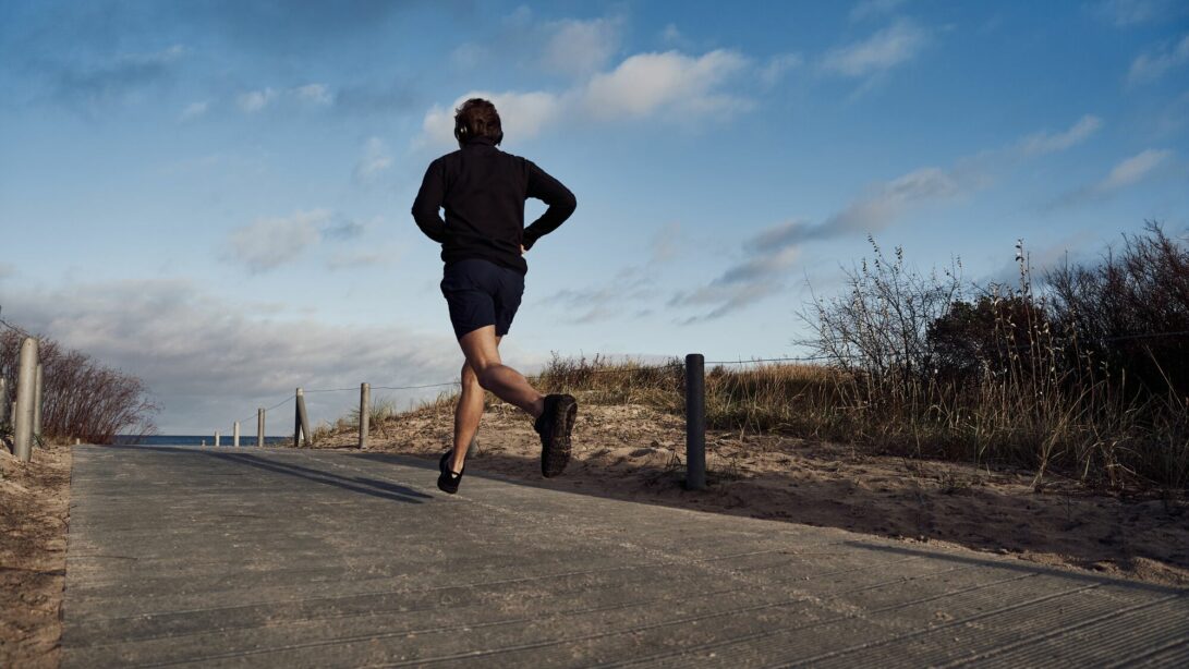 Ein Mann joggt auf einem leeren Weg in Richtung Meer. Im Hintergrund ist ein leicht bewölkter blauer Himmel.