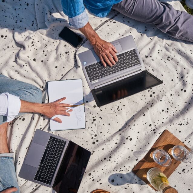 Zwei Personen im Freien auf einer Picknickdecke mit Laptops und Notizbuch
