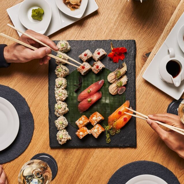 Sushi und Sashimi auf einer Schieferplatte, welches auf einem Holztisch steht. Zwei Hände greifen mit Stäbchen zum Essen