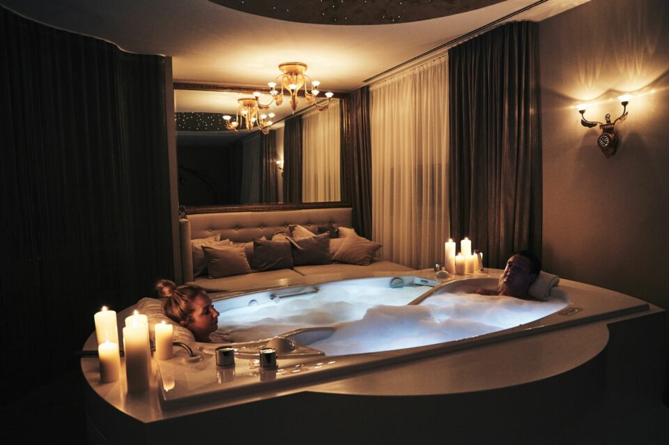 Ein Pärchen sitzt in einer Badewanne mit viel Schaum. Am Rand der Wanner stehen entzündete Kerzen.