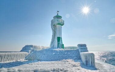 Kleiner grün-weißer Leuchtturm, welcher zur Hälfte von Eis bedeckt ist ebenso wie sein Zugang. Die Sonne scheint.