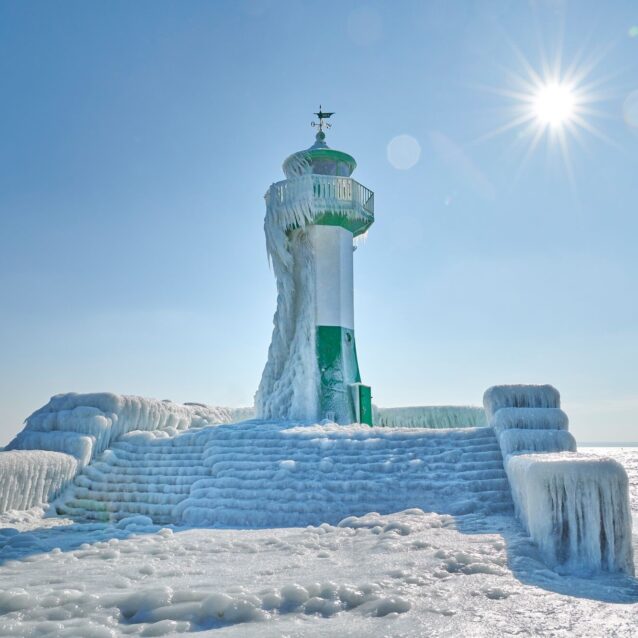 Kleiner grün-weißer Leuchtturm, welcher zur Hälfte von Eis bedeckt ist ebenso wie sein Zugang. Die Sonne scheint.