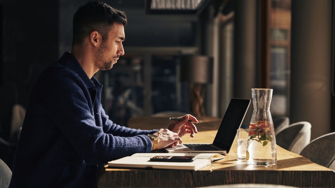 Ein Mann in einem blauen Cardigan sitzt am Tisch und arbeitet am Laptop. Sein Telefon liegt auf dem Tisch.