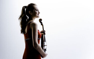 Man sieht eine junge Violinistin mit Pferdeschwanz im Profil vor einem weißen Hintergrund.. Sie hält eine Geige in den Händen und trägt ein rotes Kleid.