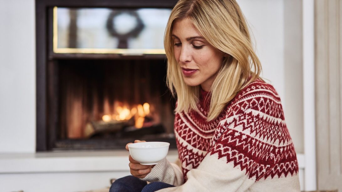 Frau in winterlichen Pullover sitzt mit einer Tasse Tee vor einem Kamin