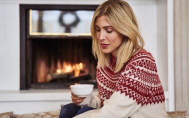 Frau in winterlichen Pullover sitzt mit einer Tasse Tee vor einem Kamin