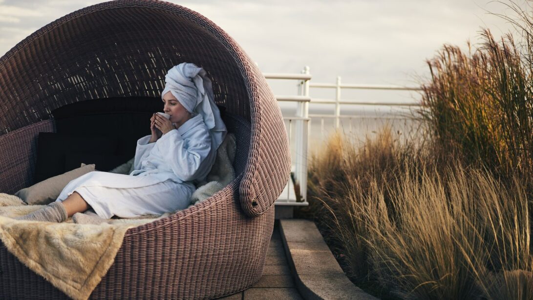 Eine Frau im Badelmantel sitzt in einem Sitzkorb auf einer Terasse vor einem Strand und trinkt aus einer Tasse.